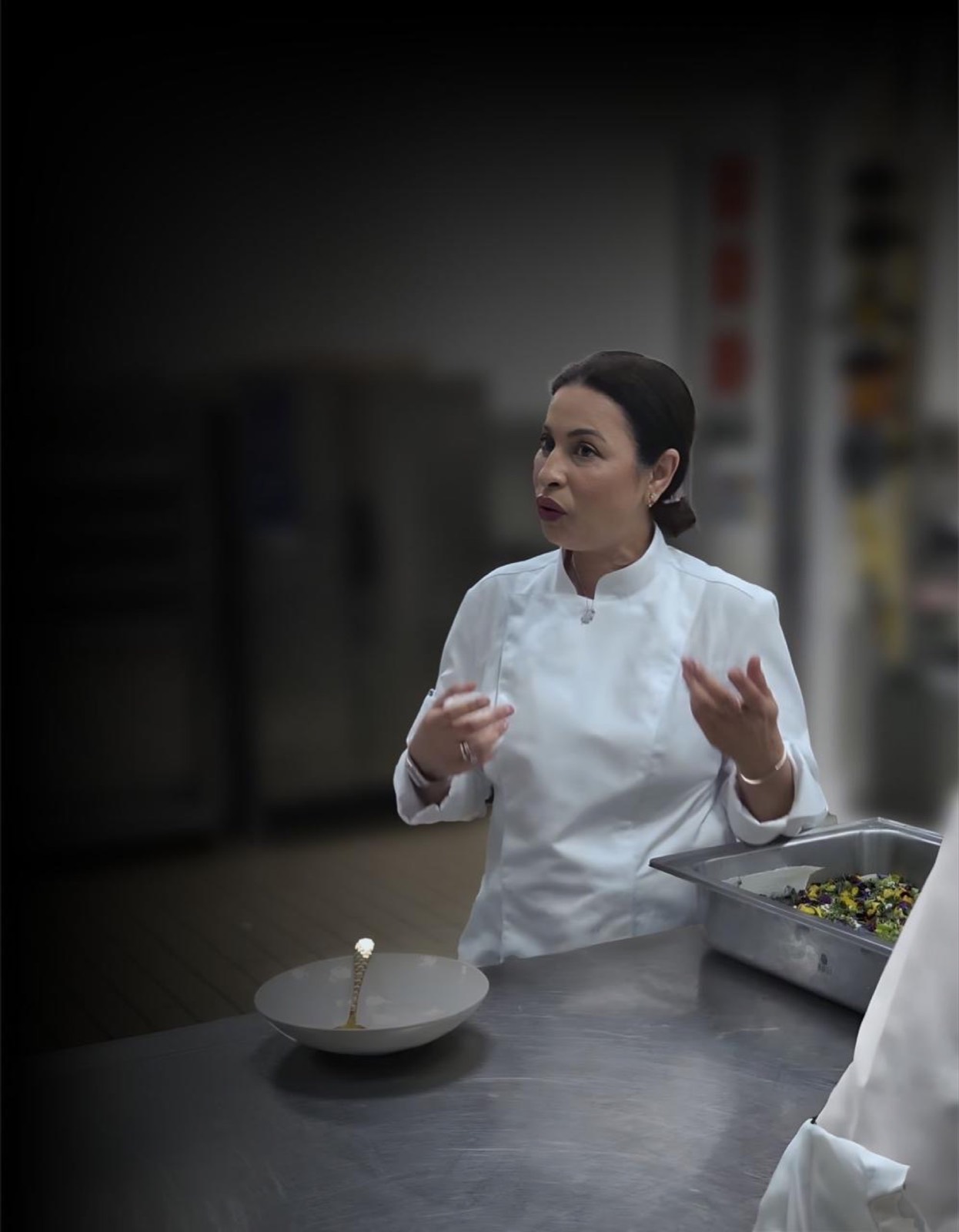 Fatima Nejjar en veste de cuisine, mains ouvertes, expliquant un dressage devant un bac de fleurs comestibles