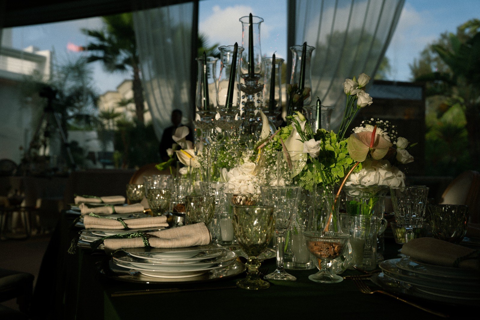 Longue table vert sombre sous voilages blancs : candélabres de cristal, fleurs blanches et vertes, lin et cristal taillé