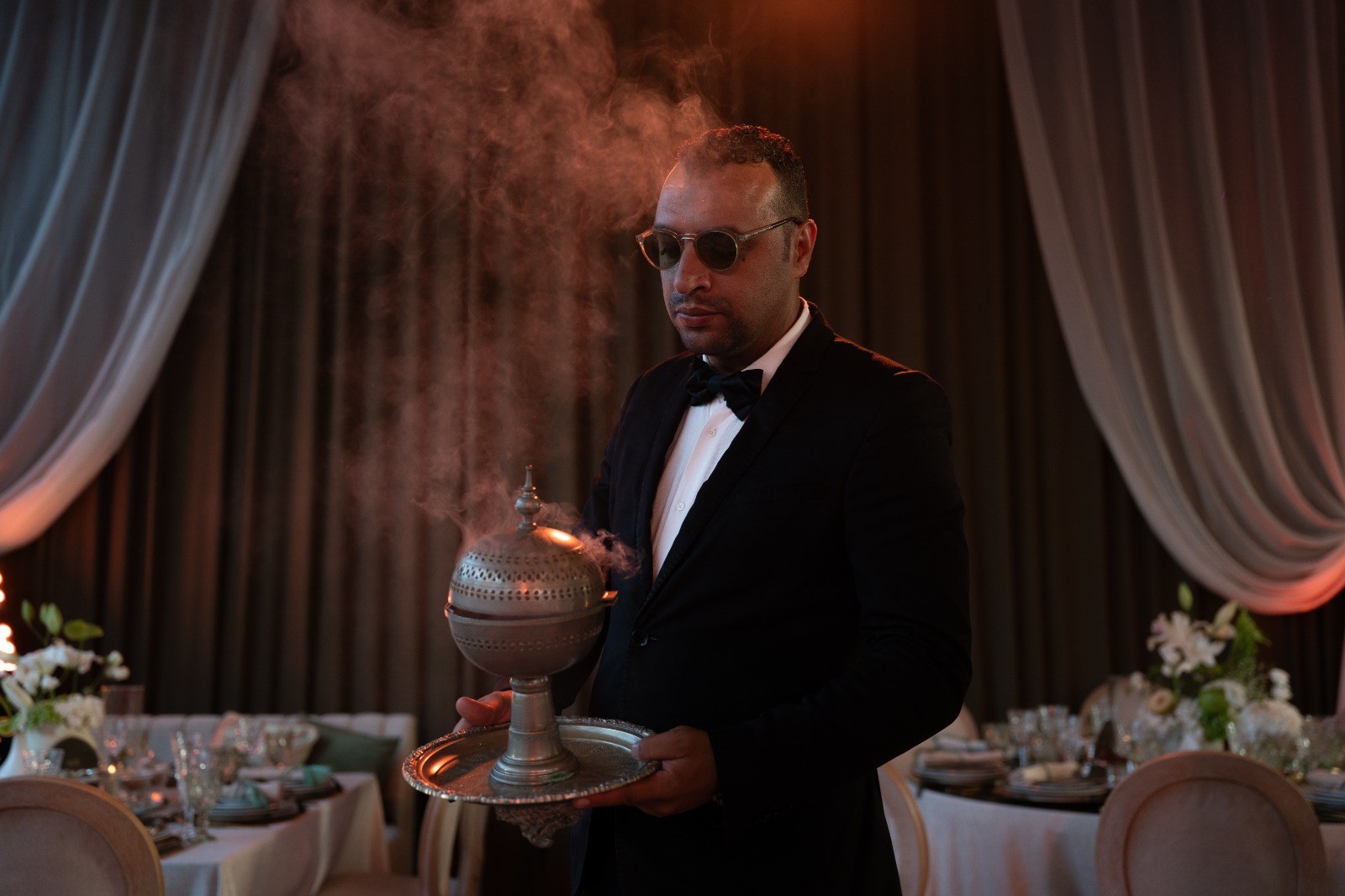 Serveur en smoking portant un brûle-parfum d'étain fumant sur un plateau d'argent, entre les tables dressées