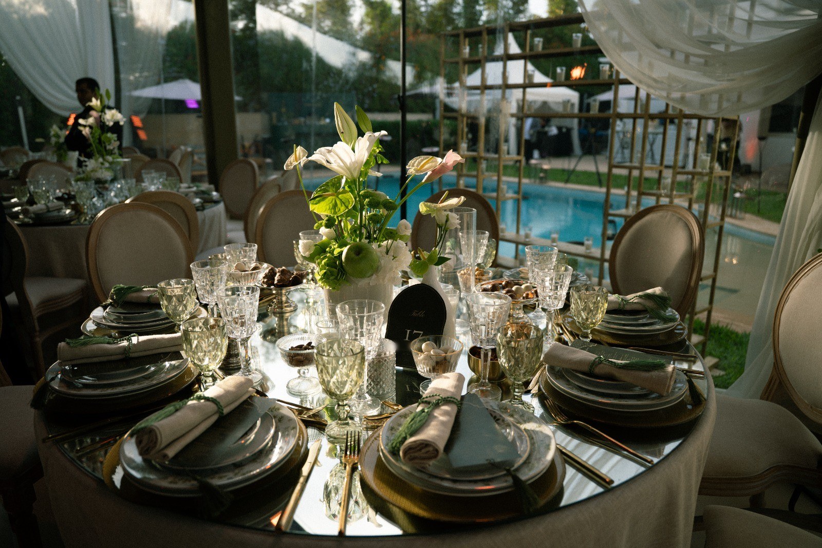 Table ronde à plateau miroir au bord d'une piscine, lys et anthuriums, cristal taillé et serviettes nouées de vert