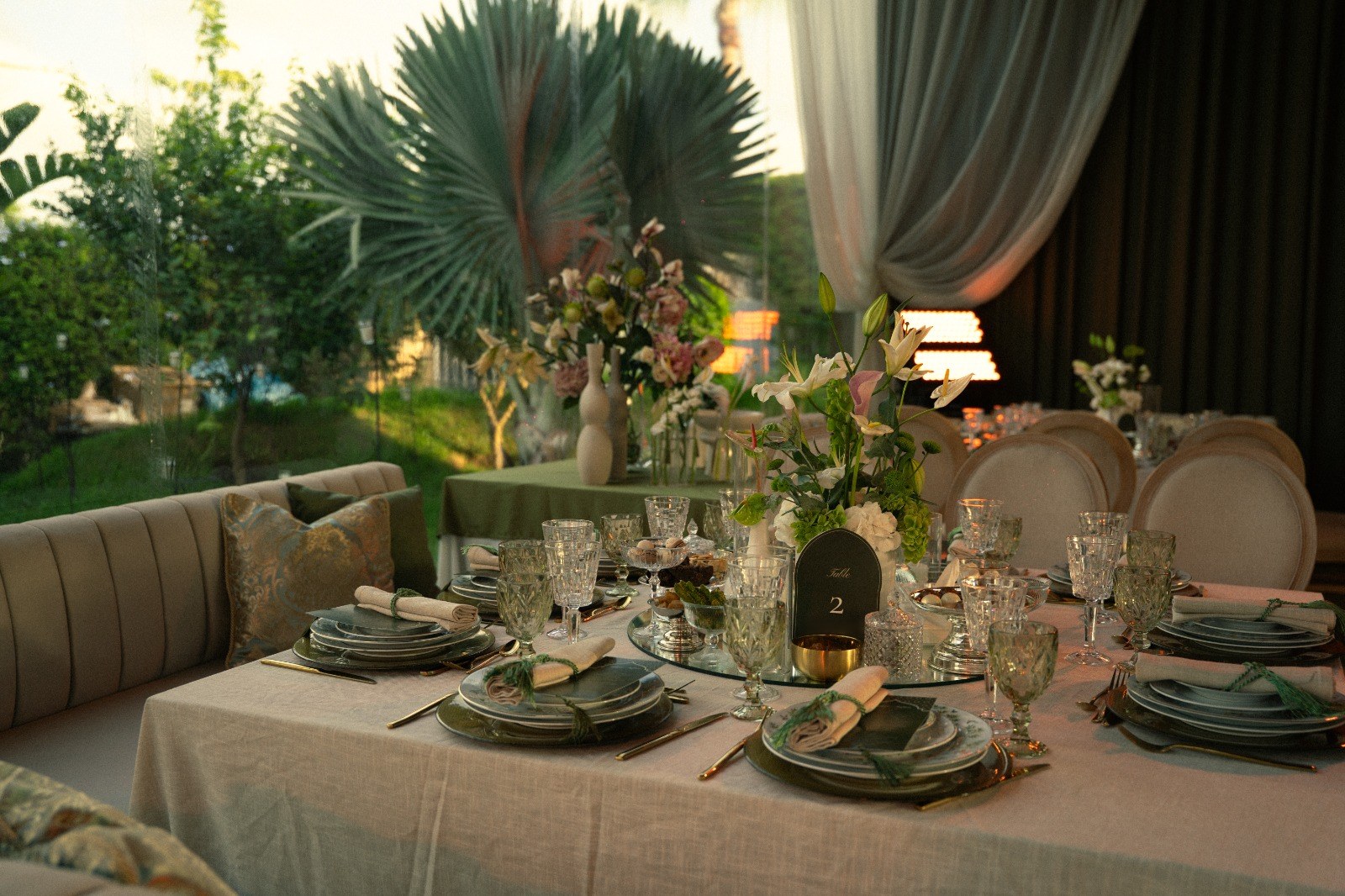 Table de lin écru le long d'une banquette à coussins verts, lys et arums, verres verts, jardin et palmier derrière