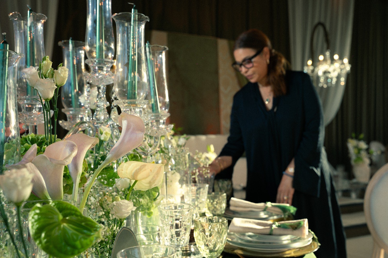 Arums et candélabres de cristal au premier plan ; derrière, Fatima Nejjar dispose les assiettes d'une table