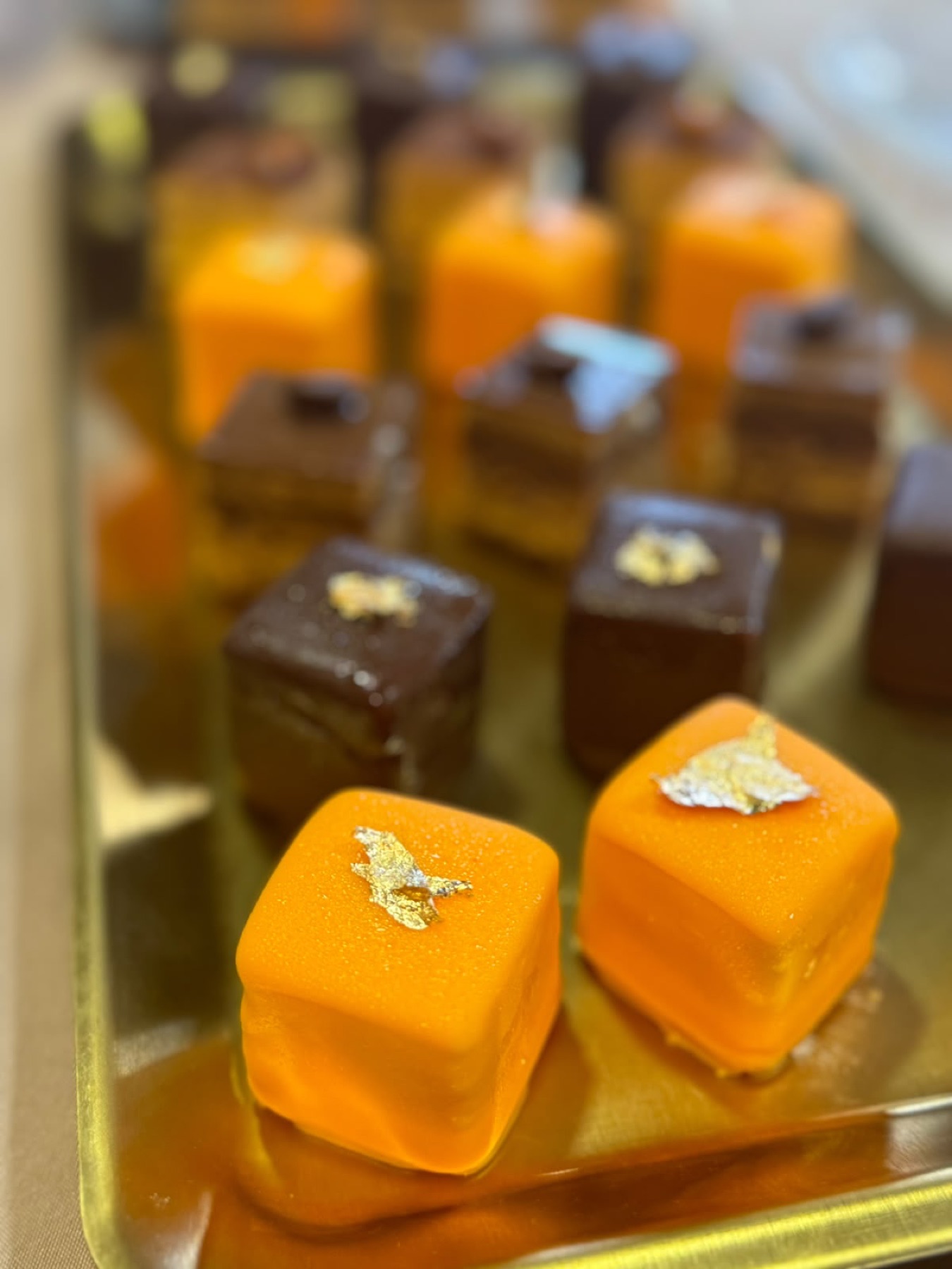 Plateau doré de cubes à glaçage orange et de carrés au chocolat, chacun marqué d'un éclat de feuille d'or