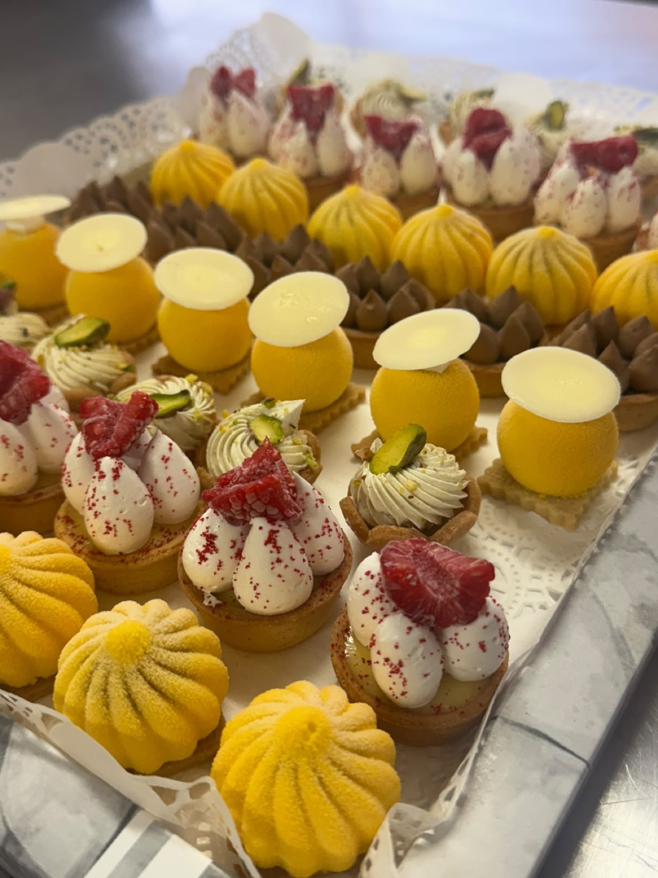 Plateau serré de mignardises : dômes et sphères jaunes floqués, tartelettes à la framboise et tartelettes pistache