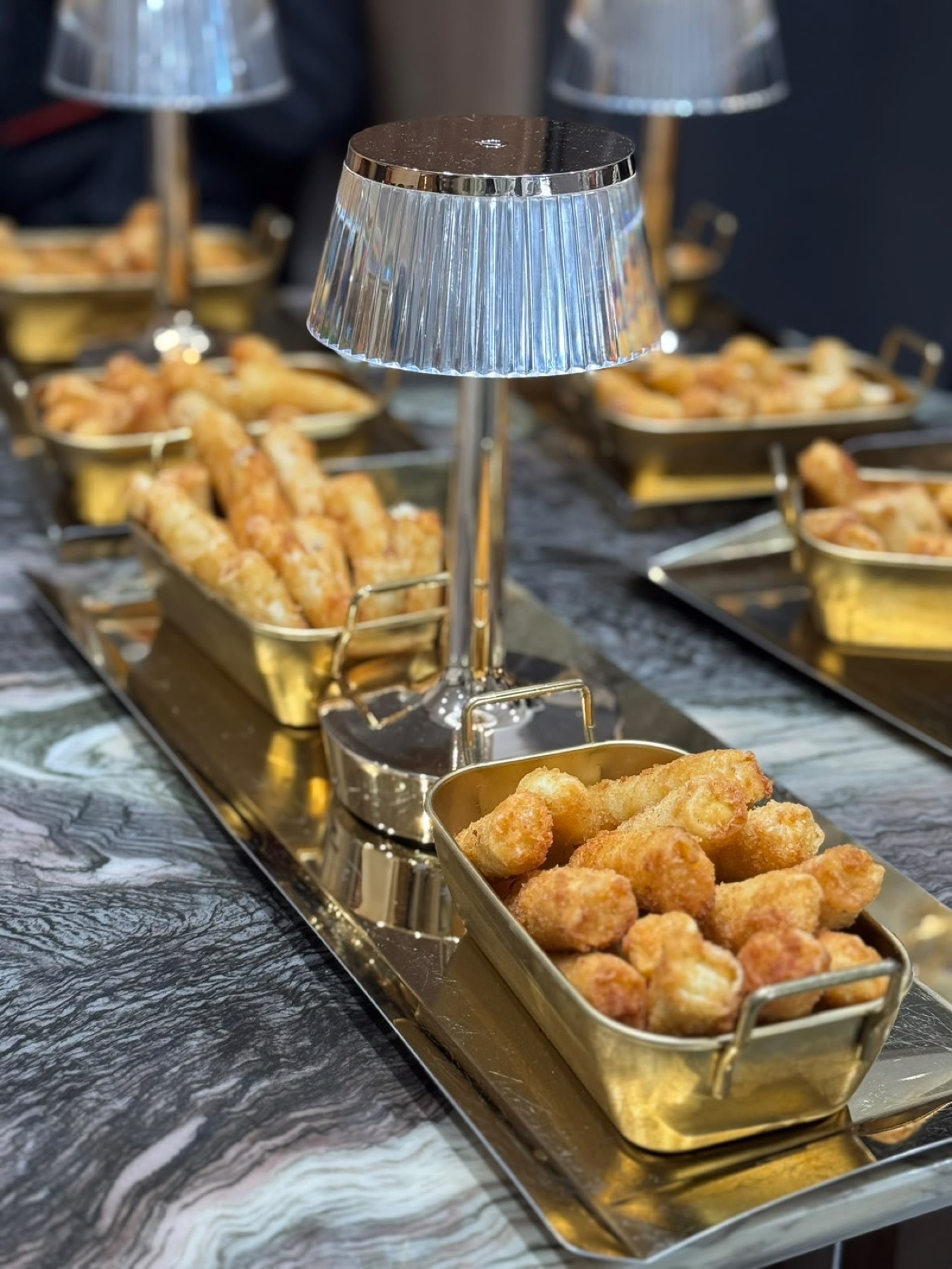 Corbeilles dorées de cigares de brick et de croquettes sur un plateau miroir, entre deux lampes de cristal